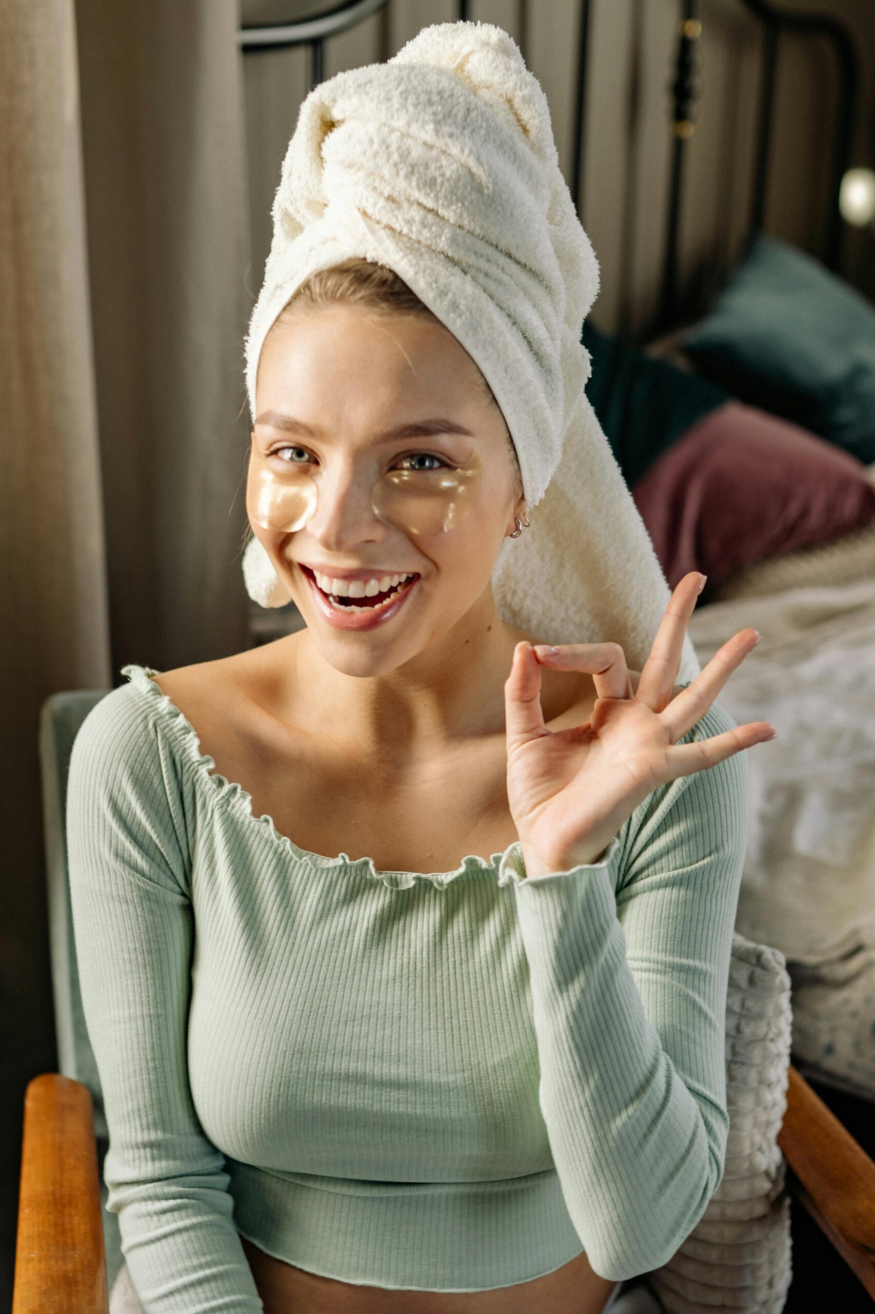 Young woman practicing skincare in home setting, smiling with eye patches and towel on head.
